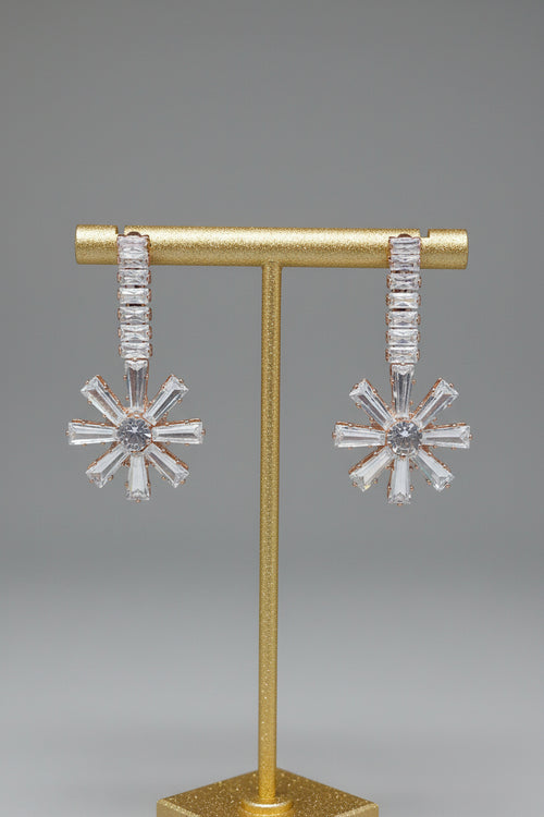 A pair of silver-toned dangle earrings with baguette-cut clear stones, displayed on a gold T-stand against a neutral gray background.