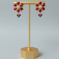 A pair of gold-toned floral drop earrings with red gemstone petals, white stones in the center, and small green bead drops, displayed on a gold stand against a light gray background.