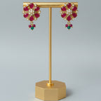A pair of gold-toned floral drop earrings with red gemstone petals, white stones in the center, and small green bead drops, displayed on a gold stand against a light gray background.