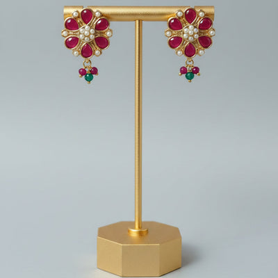 A pair of gold-toned floral drop earrings with red gemstone petals, white stones in the center, and small green bead drops, displayed on a gold stand against a light gray background.
