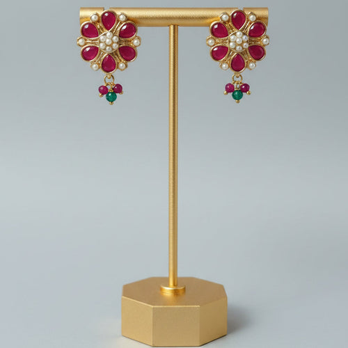 A pair of gold-toned floral drop earrings with red gemstone petals, white stones in the center, and small green bead drops, displayed on a gold stand against a light gray background.