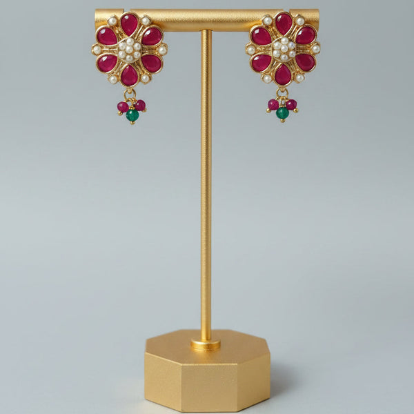 A pair of gold-toned floral drop earrings with red gemstone petals, white stones in the center, and small green bead drops, displayed on a gold stand against a light gray background.