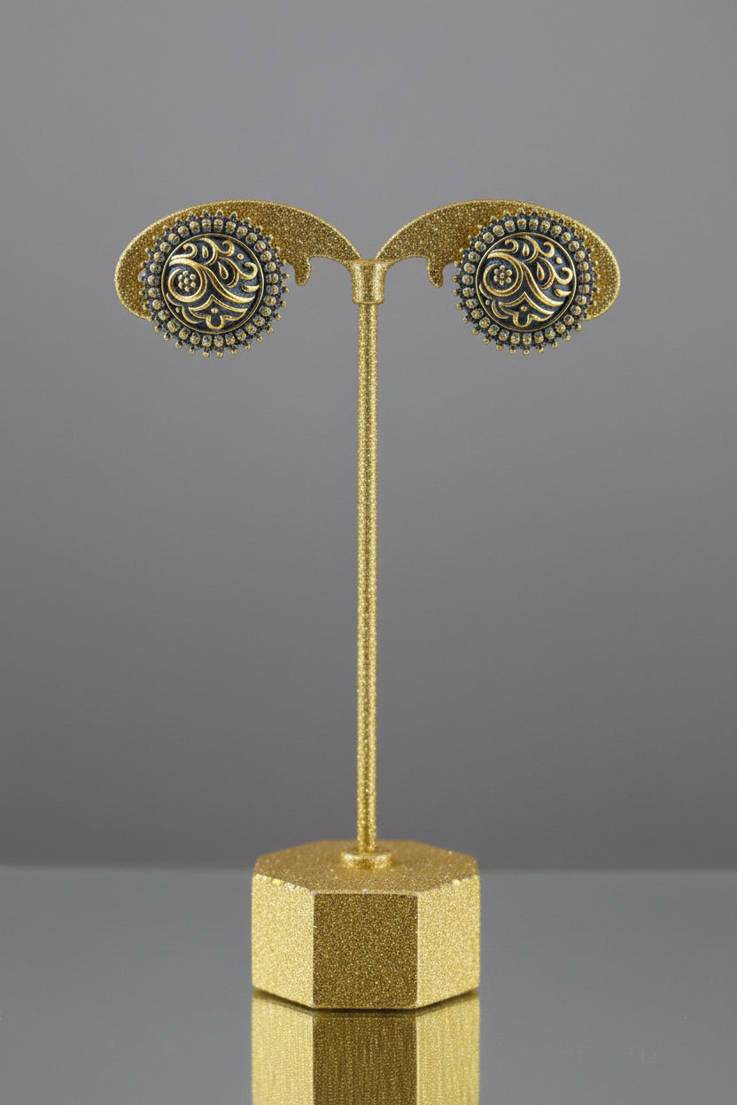 A pair of round gold and blue enamel earrings displayed on a black stand attached to a light wood block, set against a red and beige background.