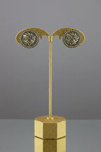 A pair of round gold and blue enamel earrings displayed on a black stand attached to a light wood block, set against a red and beige background.