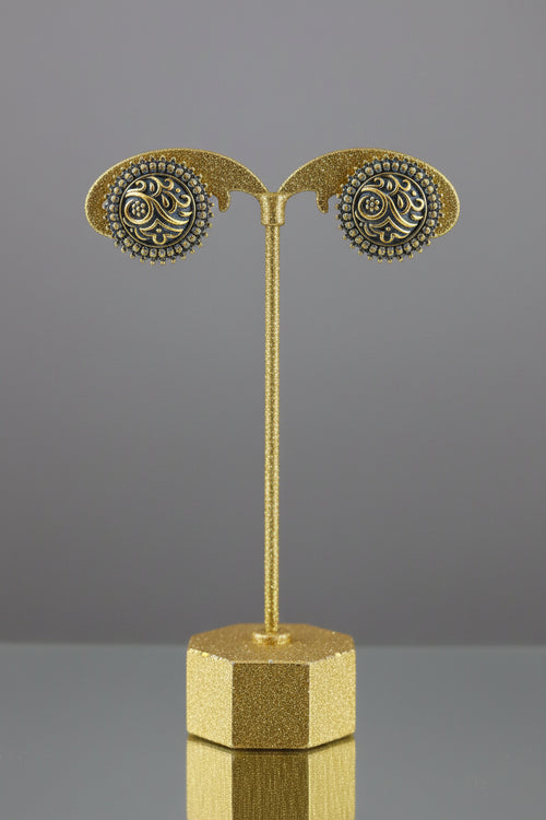 A pair of round gold and blue enamel earrings displayed on a black stand attached to a light wood block, set against a red and beige background.