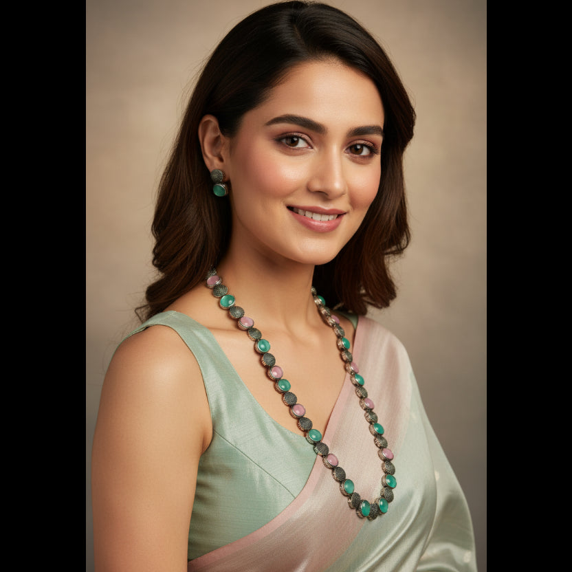 A jewelry set featuring a necklace and matching earrings with round green and pink stones set in silver-toned metal, displayed on a clear plastic stand.