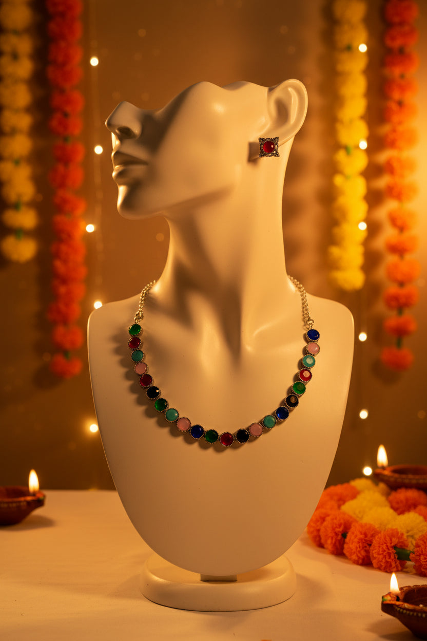 A silver-tone necklace and matching stud earrings set with round, colorful gemstone-like beads in red, green, pink, blue, black, and white, displayed on a light fabric background.