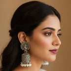 A woman in profile wearing ornate silver-toned jhumka earrings with white pearl drops, against a warm beige background.