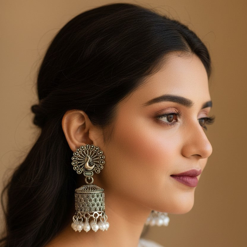 A woman in profile wearing ornate silver-toned jhumka earrings with white pearl drops, against a warm beige background.