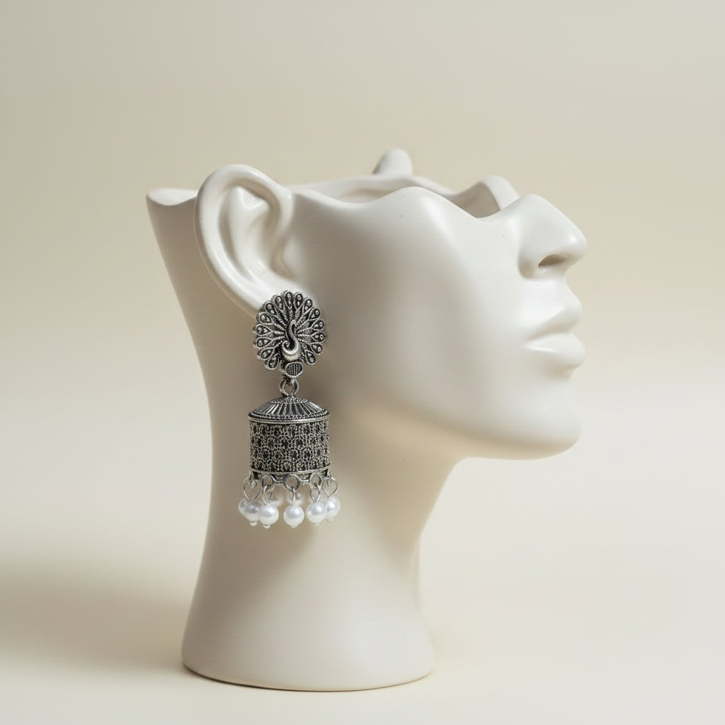 A pair of silver-toned traditional Indian jhumka earrings with peacock head motifs and white pearl bead drops, displayed on a black stand with a white card labeled (NG).