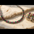 A silver-toned jewelry set featuring a necklace and matching earrings with oval green and round pink stones, displayed on a light beige stone surface.