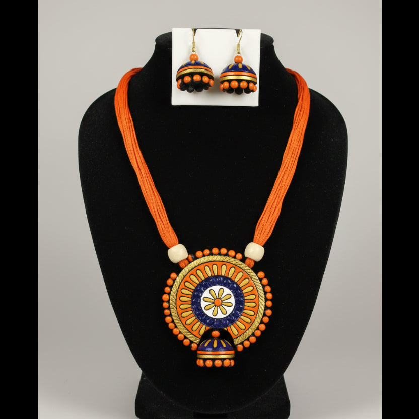 A traditional Indian jewelry set featuring a necklace and matching earrings displayed on a white mannequin bust. The jewelry has vibrant orange cords, blue and gold painted jhumka-style pendants, and orange bead details.