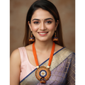 A smiling woman wearing a traditional Indian outfit with a lavender and gold saree, a light pink blouse, and a prominent orange and gold statement necklace and matching earrings, set against a warm brown studio background.