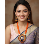 A smiling woman wearing a traditional Indian outfit with a lavender and gold saree, a light pink blouse, and a prominent orange and gold statement necklace and matching earrings, set against a warm brown studio background.