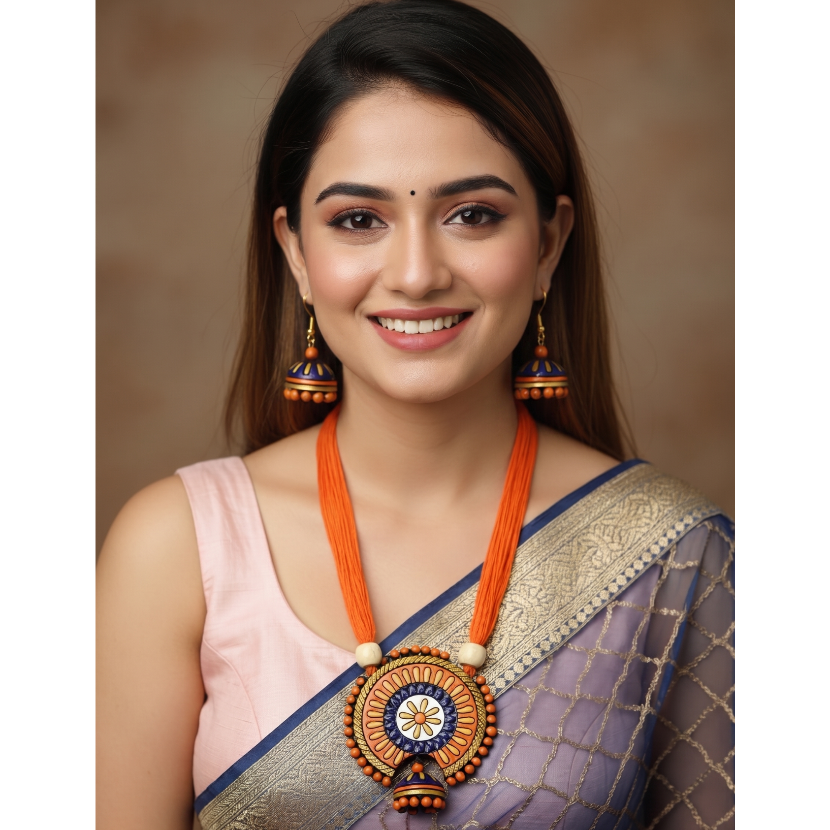 A smiling woman wearing a traditional Indian outfit with a lavender and gold saree, a light pink blouse, and a prominent orange and gold statement necklace and matching earrings, set against a warm brown studio background.