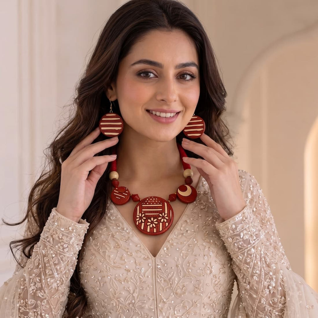 A woman wearing a red and beige traditional necklace and matching earrings, holding them up to show the details of the jewelry against a soft beige background.