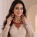A woman wearing a red and beige traditional necklace and matching earrings, holding them up to show the details of the jewelry against a soft beige background.