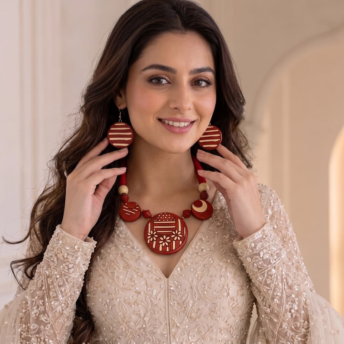 A woman wearing a red and beige traditional necklace and matching earrings, holding them up to show the details of the jewelry against a soft beige background.