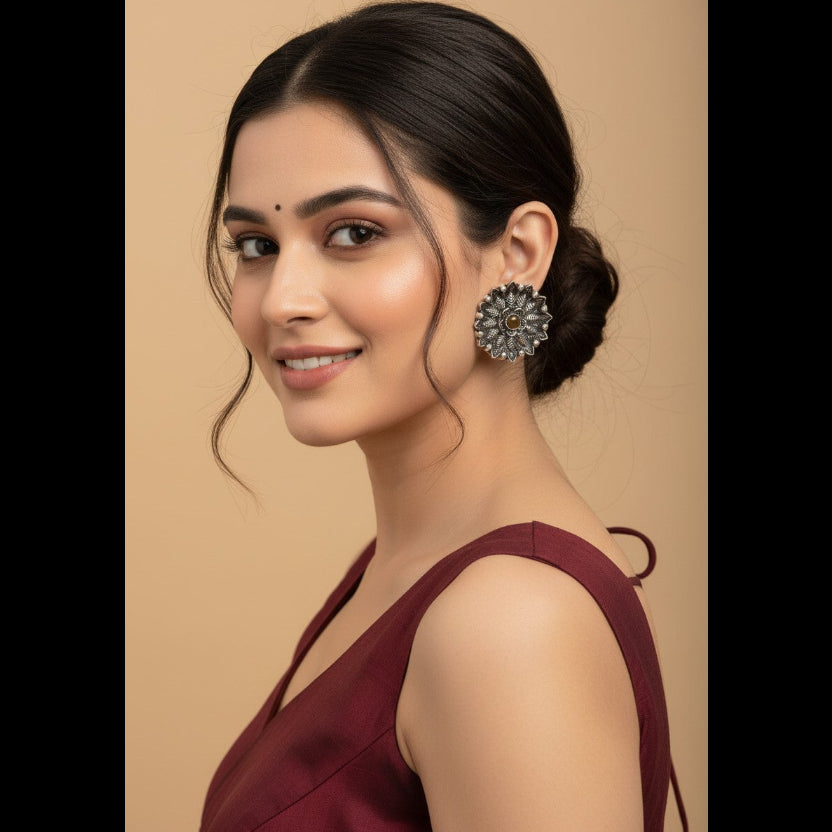 A woman with dark hair in a low bun wears a large, ornate silver-toned floral earring with clear stones, set against a solid beige background.