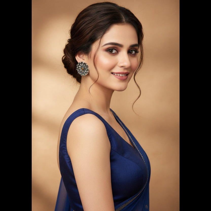 A woman in a deep blue sleeveless dress with a sheer draped neckline, wearing a large silver-toned floral earring, set against a warm beige background.