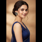 A woman in a deep blue sleeveless dress with a sheer draped neckline, wearing a large silver-toned floral earring, set against a warm beige background.