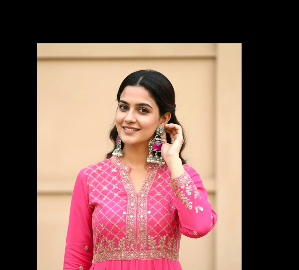 A woman wearing a bright pink traditional outfit with gold embroidery and large dangling earrings, standing against a beige wall.