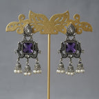 A pair of ornate silver-toned dangle earrings with square purple gemstones and small pearl-like drops, displayed on a gold stand against a soft gray background.