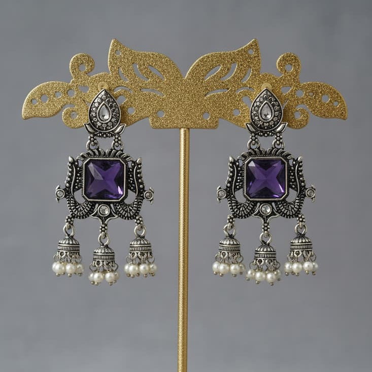 A pair of ornate silver-toned dangle earrings with square purple gemstones and small pearl-like drops, displayed on a gold stand against a soft gray background.