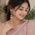 A woman wearing ornate silver-toned jhumka earrings with white pearl drops, photographed in a softly lit indoor setting with blurred greenery in the background.