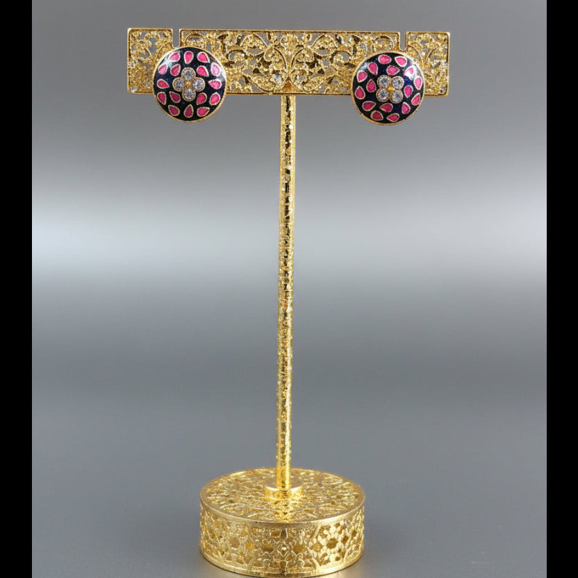 A pair of round stud earrings displayed on a small white card stand, resting on a wooden block over a red surface. The earrings have a gold-tone setting with a pink and red floral pattern and clear rhinestones in the center.