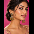 A pair of ornate gold-toned jhumka earrings with pink and white stone embellishments, hanging from a white jewelry display card.