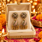 A pair of ornate gold-toned jhumka earrings with pink and white stone embellishments, hanging from a white jewelry display card.