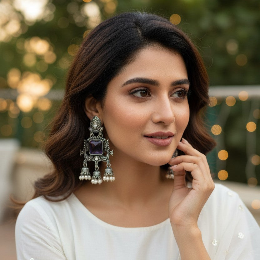 A woman wearing ornate silver-toned traditional earrings with dark purple rectangular stones and white pearl-like drops, styled with a white embroidered top against a softly blurred outdoor background.