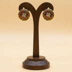 A pair of round gold-tone earrings with red, green, and white floral enamel detailing, displayed on a dark wood T-stand against a warm beige background.