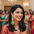 A smiling woman in a traditional Indian outfit with long, ornate gold and red beaded earrings, standing in a grand hall with blurred people in the background.