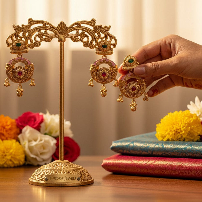A hand wearing gold bangles holds a gold-toned traditional Indian earring next to a matching earring displayed on an ornate gold stand. The scene is styled with folded red and blue silk fabrics and colorful flowers on a wooden surface against a warm beige background.