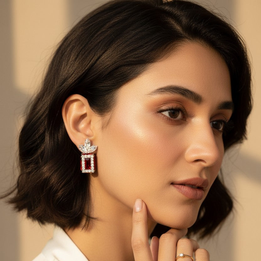 A woman with short dark hair wearing a dangling rectangular earring with a silver-toned frame and pink gemstones, paired with a gold ring on her finger. She is wearing a white top against a warm beige background with soft shadows.