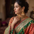 A woman in profile wearing an ornate red and green embroidered traditional garment with gold zari detailing, paired with matching gold jhumka earrings, in a softly blurred indoor setting.