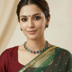 A woman wearing a silver-toned necklace and matching earrings with circular red and green stone accents, paired with a deep maroon top against a soft beige background.