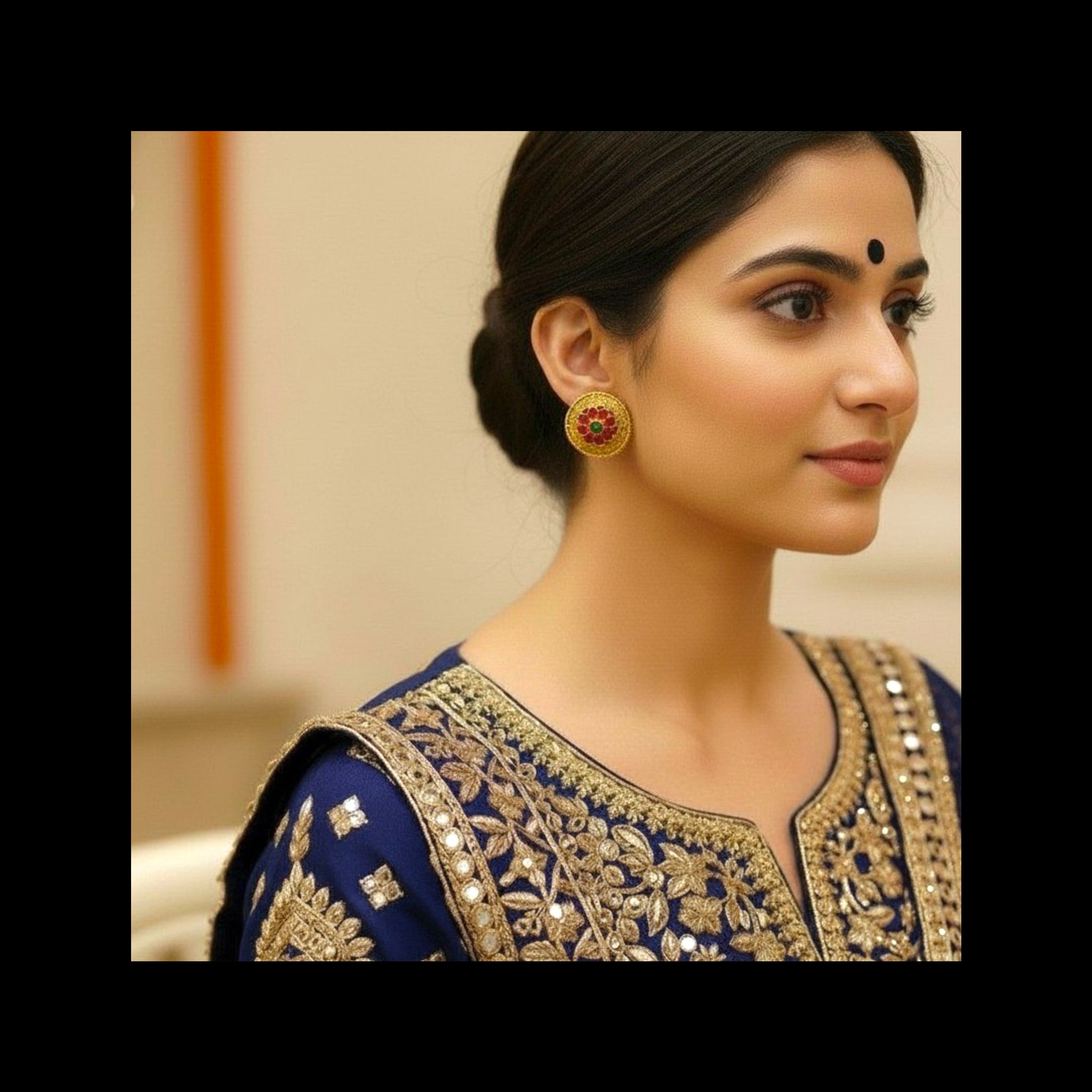 A woman in profile wearing a navy blue traditional outfit with ornate gold embroidery and a matching gold earring, photographed indoors against a softly blurred beige background.
