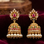 A pair of ornate gold-toned Jhumka earrings with red and green stone accents and white pearl bead drops, displayed on a deep burgundy velvet fabric.
