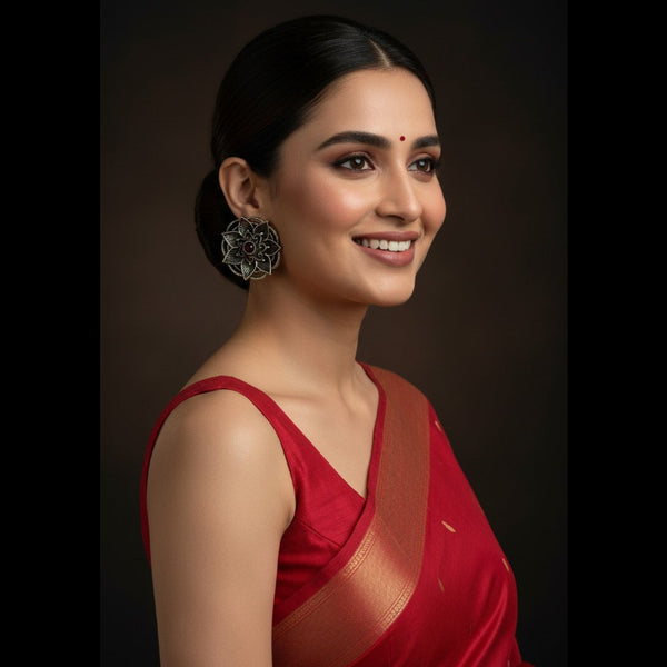 A woman in a red saree with a gold border and silver floral earrings, shown in a close-up portrait against a dark brown background.