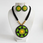 A handmade jewelry set featuring a necklace and matching earrings with circular green and yellow painted medallions on a black cord, displayed on a white bust against a green artificial grass background.