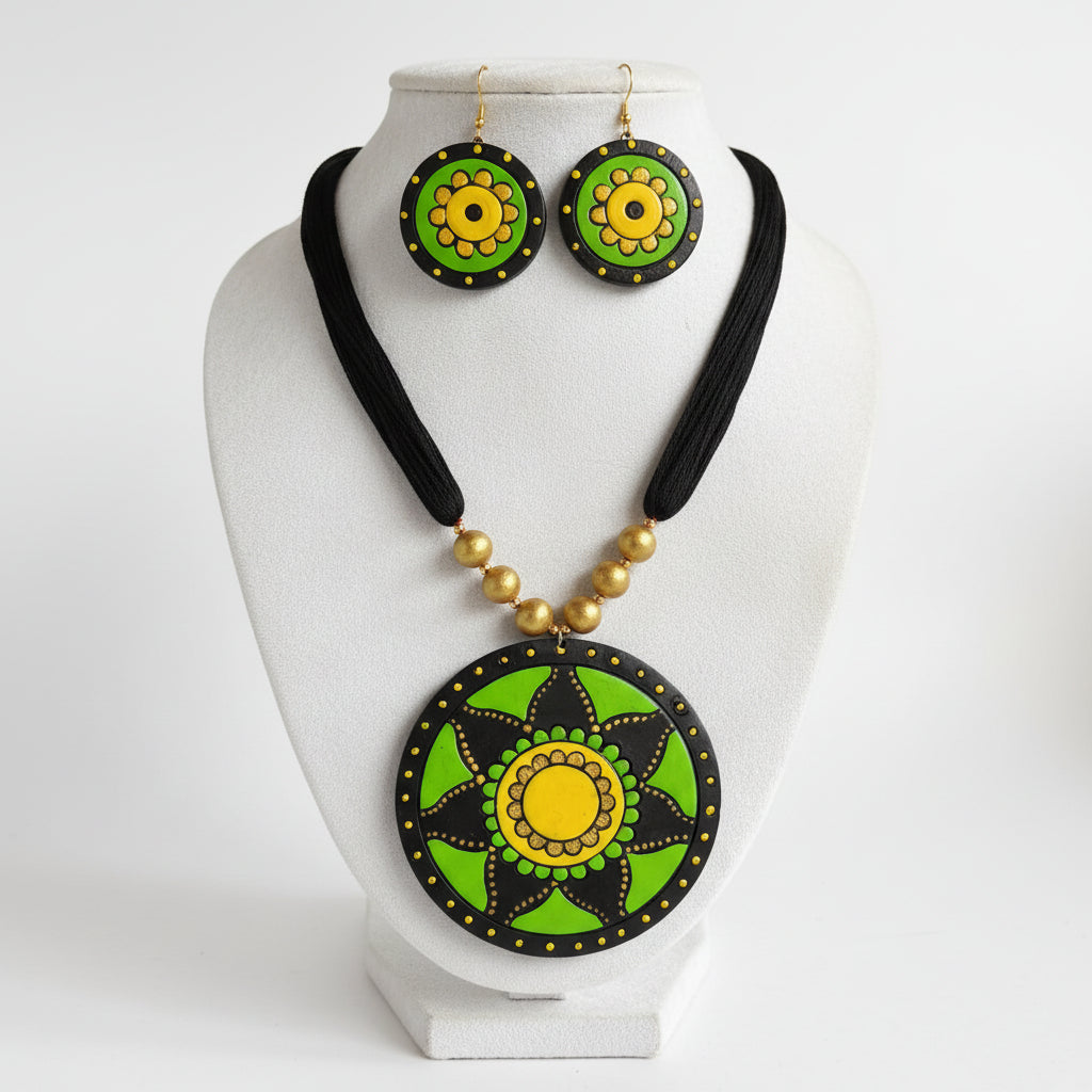 A handmade jewelry set featuring a necklace and matching earrings with circular green and yellow painted medallions on a black cord, displayed on a white bust against a green artificial grass background.