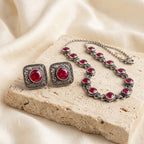 Silver-tone jewelry set with ornate filigree detailing and round red gemstones, including a necklace and matching earrings, arranged on a beige stone slab with draped cream fabric in the background.