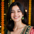A smiling woman with dark shoulder-length hair wearing a dark green embroidered traditional top with gold detailing and a red dupatta with gold borders, accessorized with green jhumka earrings, set against a warm, decorated background with marigold garlands.