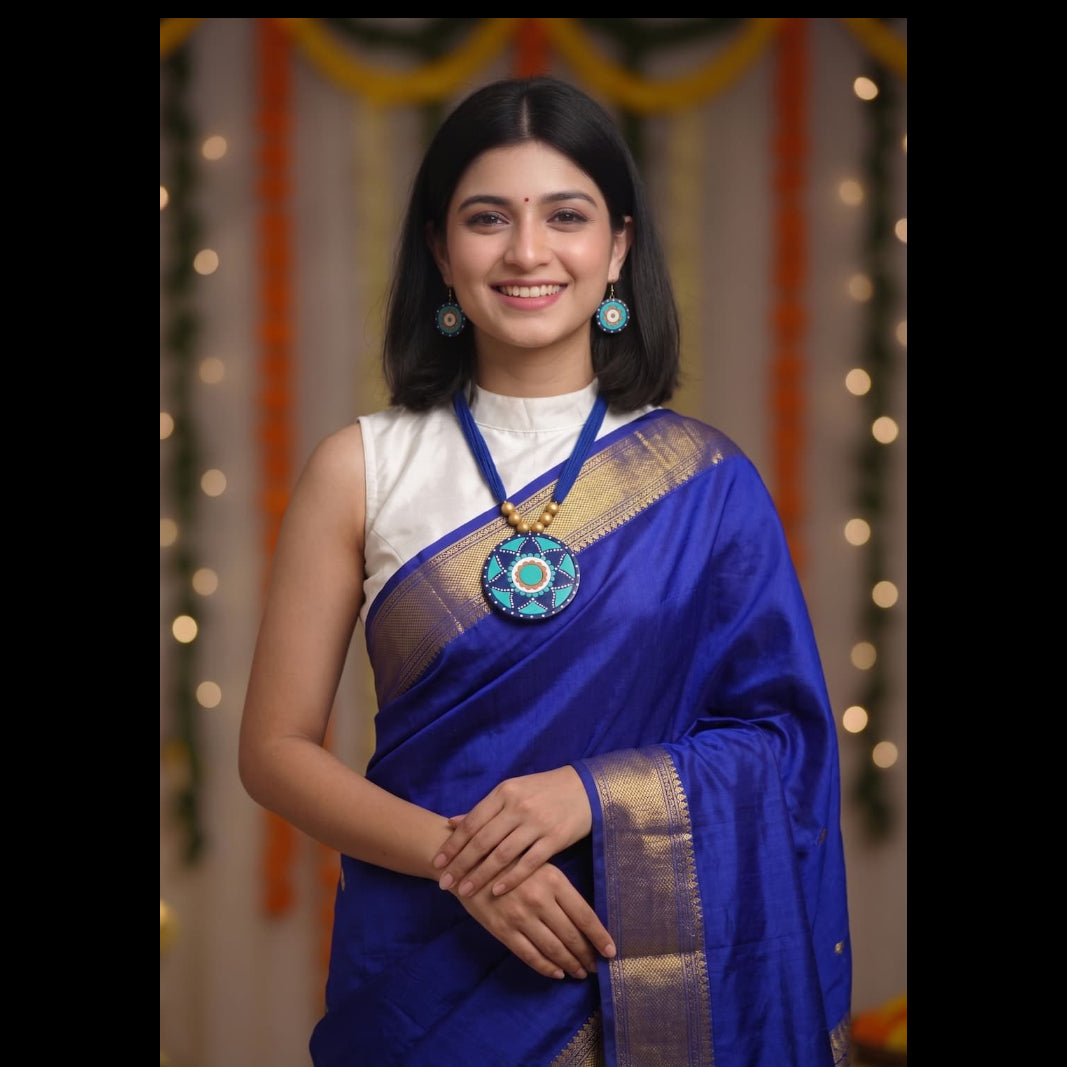 A woman wearing a royal blue silk saree with gold zari borders, paired with a white sleeveless blouse and a large circular blue and gold pendant necklace, standing against a decorated festive background with warm bokeh lights.