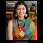 A woman with dark hair is shown from the chest up, wearing a colorful geometric beaded necklace and matching drop earrings. She is wearing a sleeveless top and a striped sari in shades of orange, teal, green, and yellow, set against a blurred indoor background.
