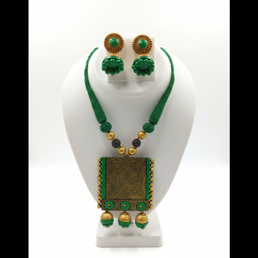 A green and gold jewelry set displayed on a white mannequin bust, featuring a necklace with a rectangular gold pendant, matching round earrings, and green beaded strands.