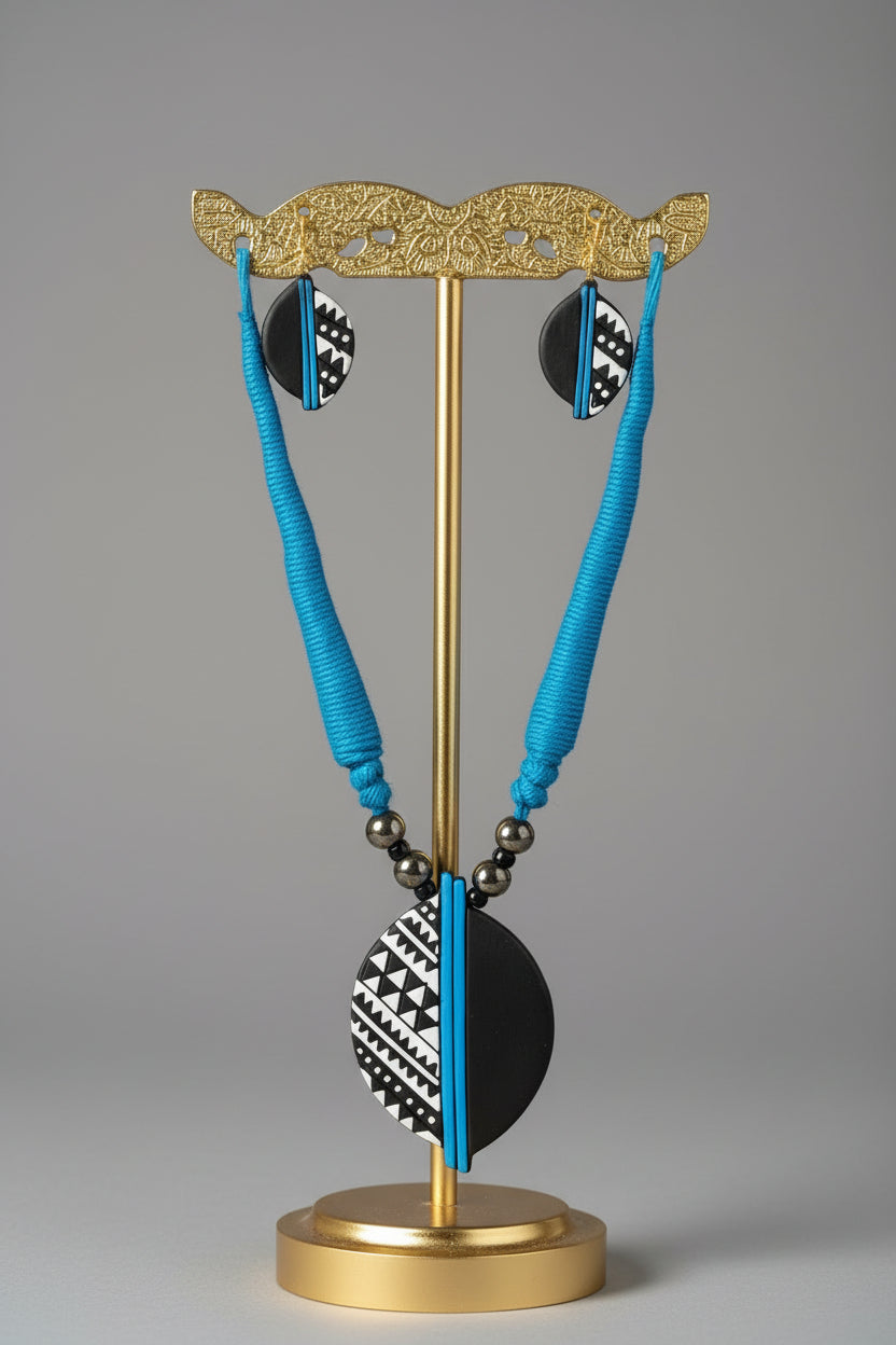 A handmade jewelry set featuring a matching necklace and earrings with black and white patterned stone discs and bright blue cords, displayed on a white card.